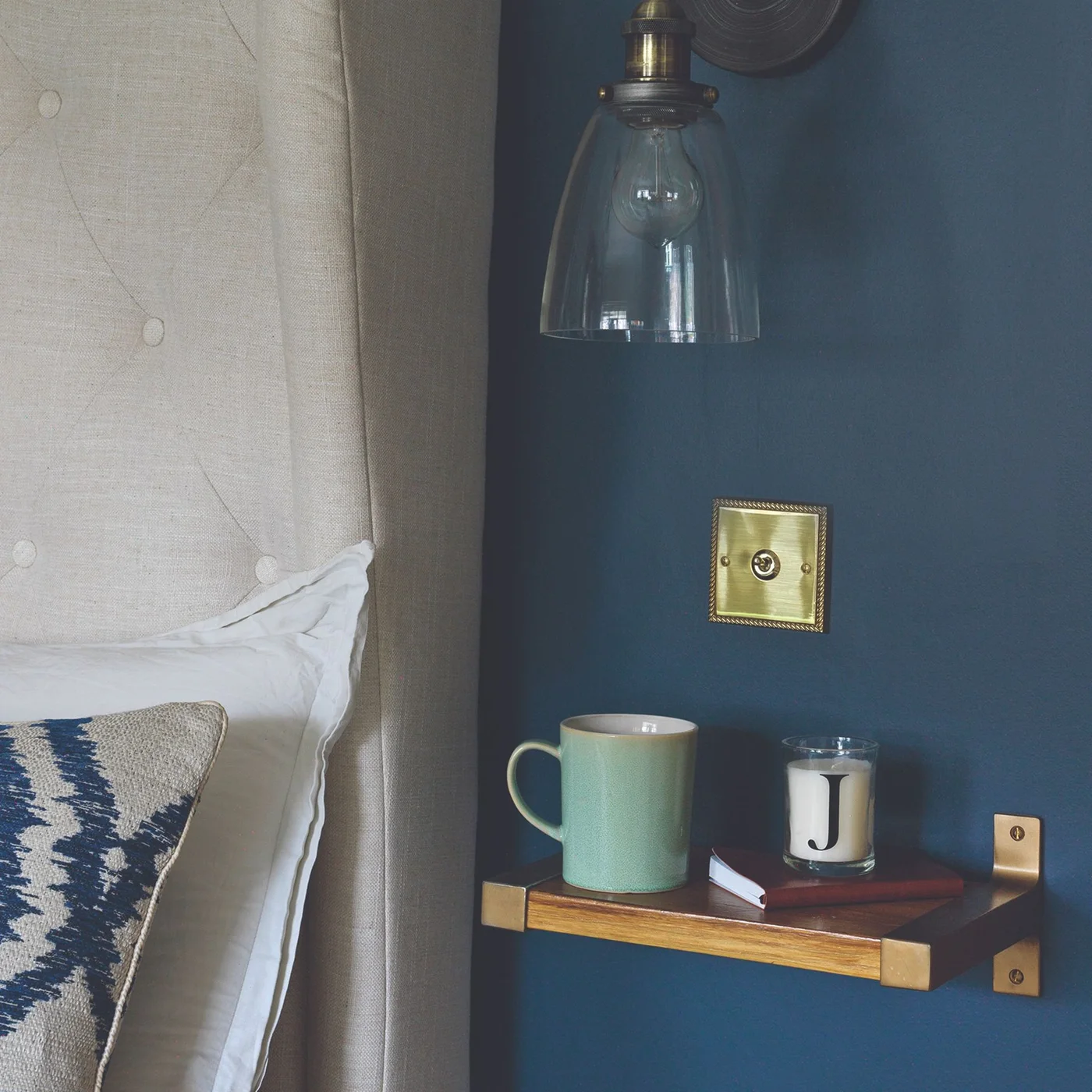 A small wood shelf with gold-colored brackets holds a mug, book and candle beside a pale upholstered headboard on a dark blue wall under a glass light.