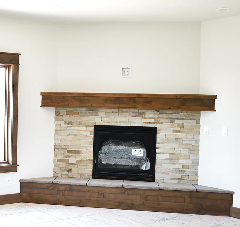 Pale textured corner fireplace with a wood mantel and raised wood-fronted hearth between windows.