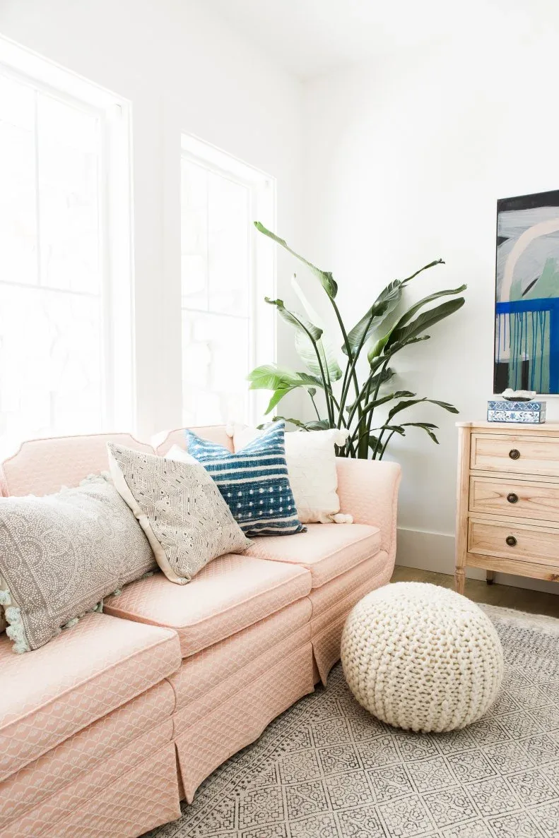 Pale pink sofa beside tall windows with a blue striped cushion, knitted pouf and wooden chest.