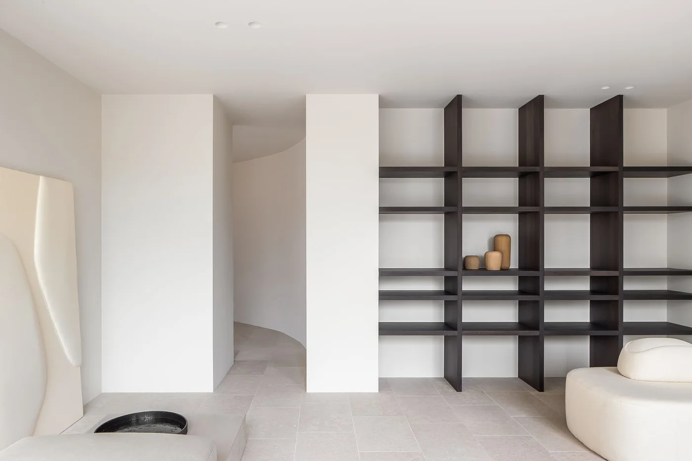 A pale apartment sitting area has a large dark grid shelf, rounded pale seat, simple wall mirror and light tiled floor