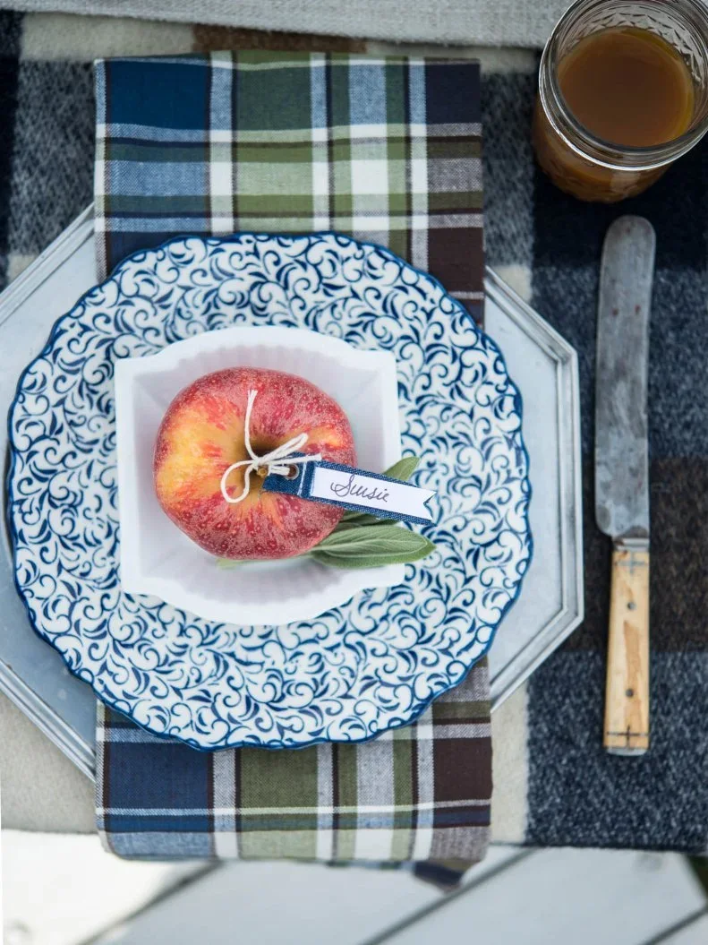 An apple with a tied name tag sits on a blue patterned plate above a checked napkin with a small green leaf