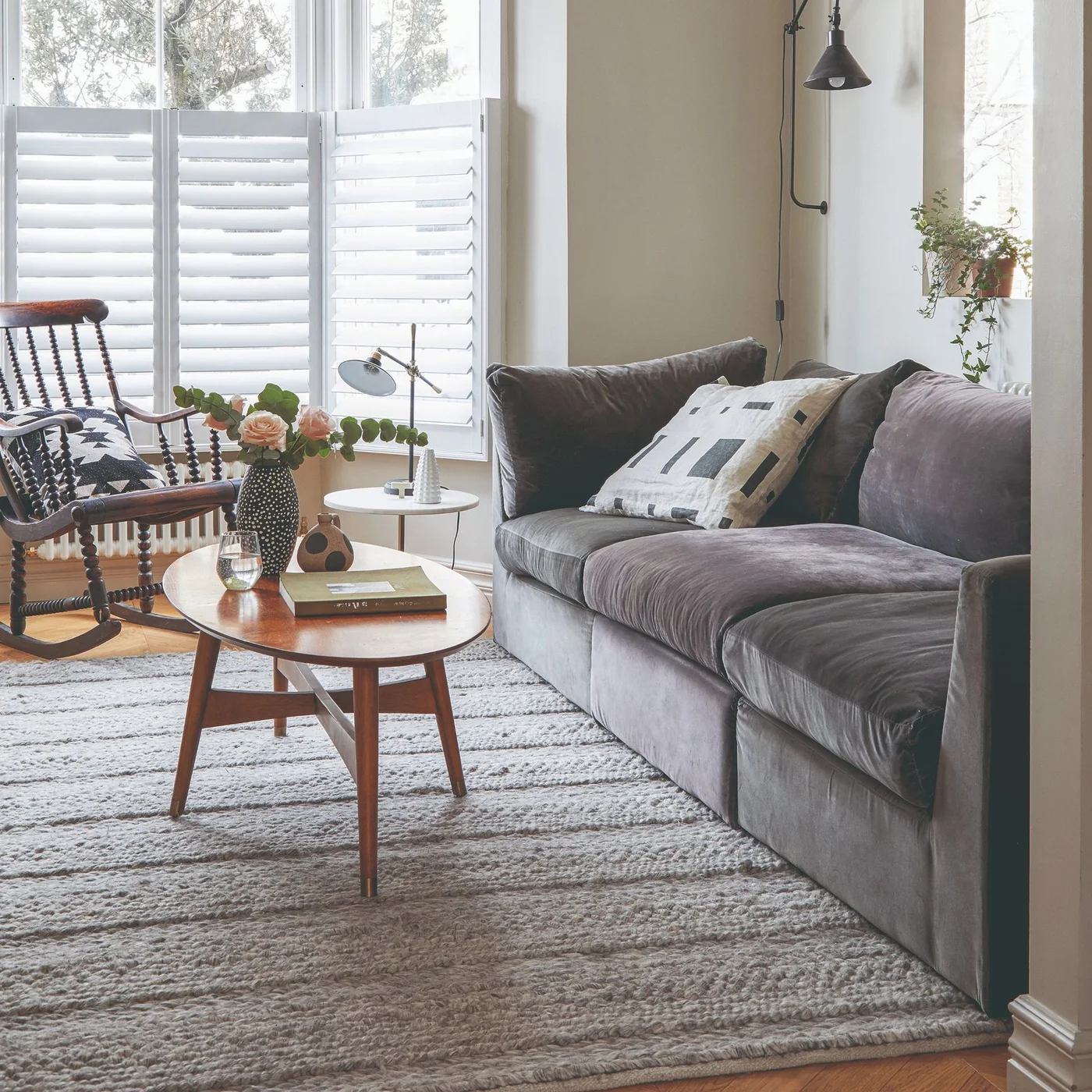A soft gray sofa sits beside white shutters and a wooden rocking chair behind a small oval wooden table on a textured pale rug