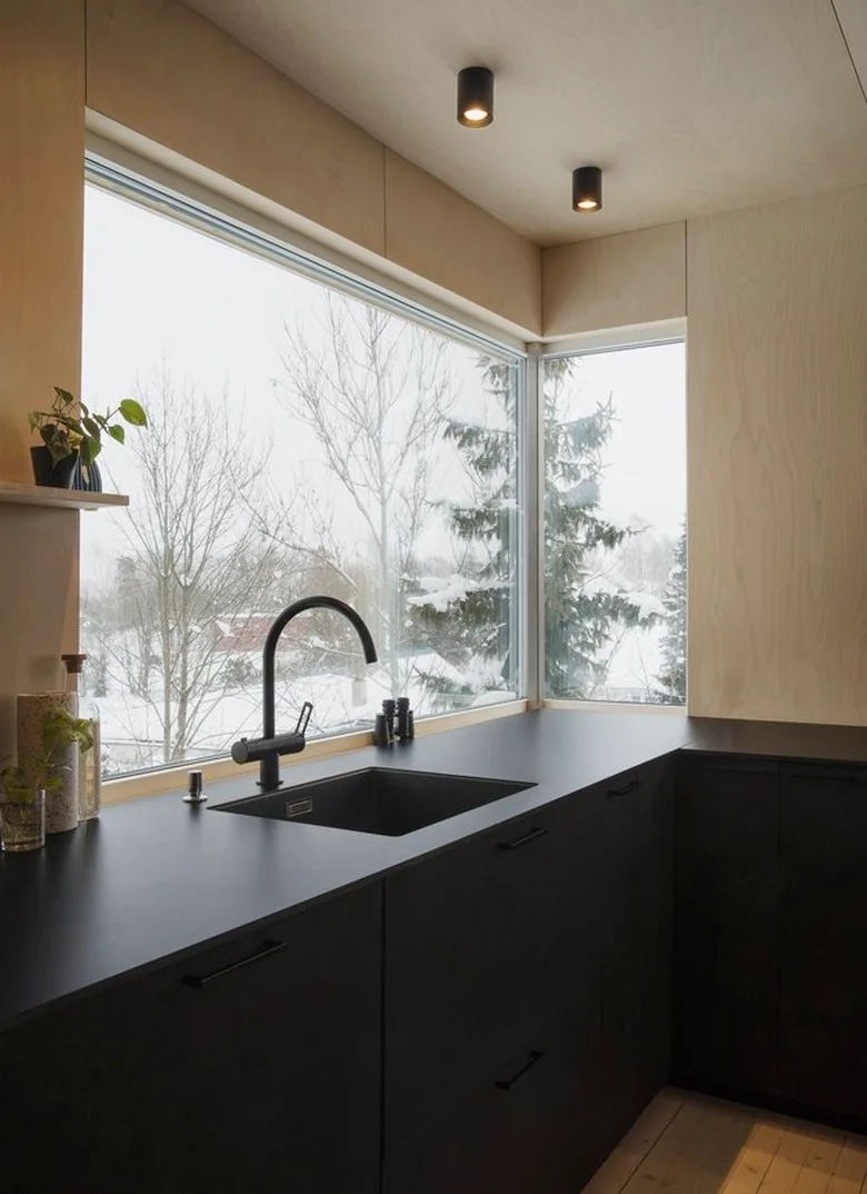 A dark kitchen sink run sits beneath a broad corner window, with pale wood-toned walls, simple black handles and small cylindrical ceiling lights.