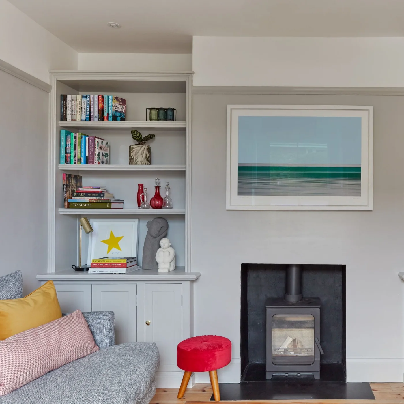 Pale gray shelves and a lower cupboard fill an alcove beside a stove, gray sofa and small red stool.