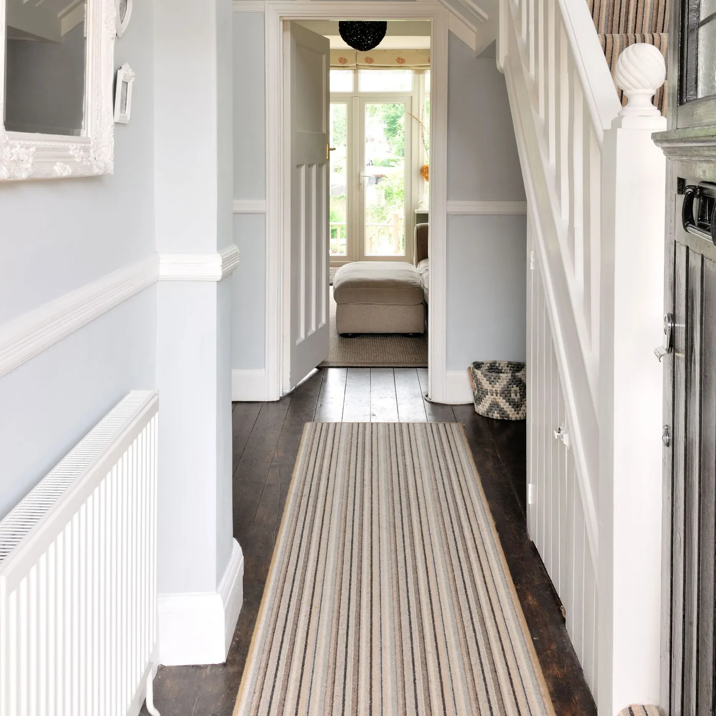 Narrow pale striped runner on dark wood floorboards beside a radiator and staircase in a light country hallway
