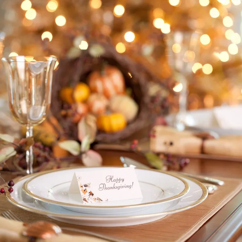 A folded Happy Thanksgiving card stands on a pale gold-rimmed plate beside clear glasses and warm-toned decorations