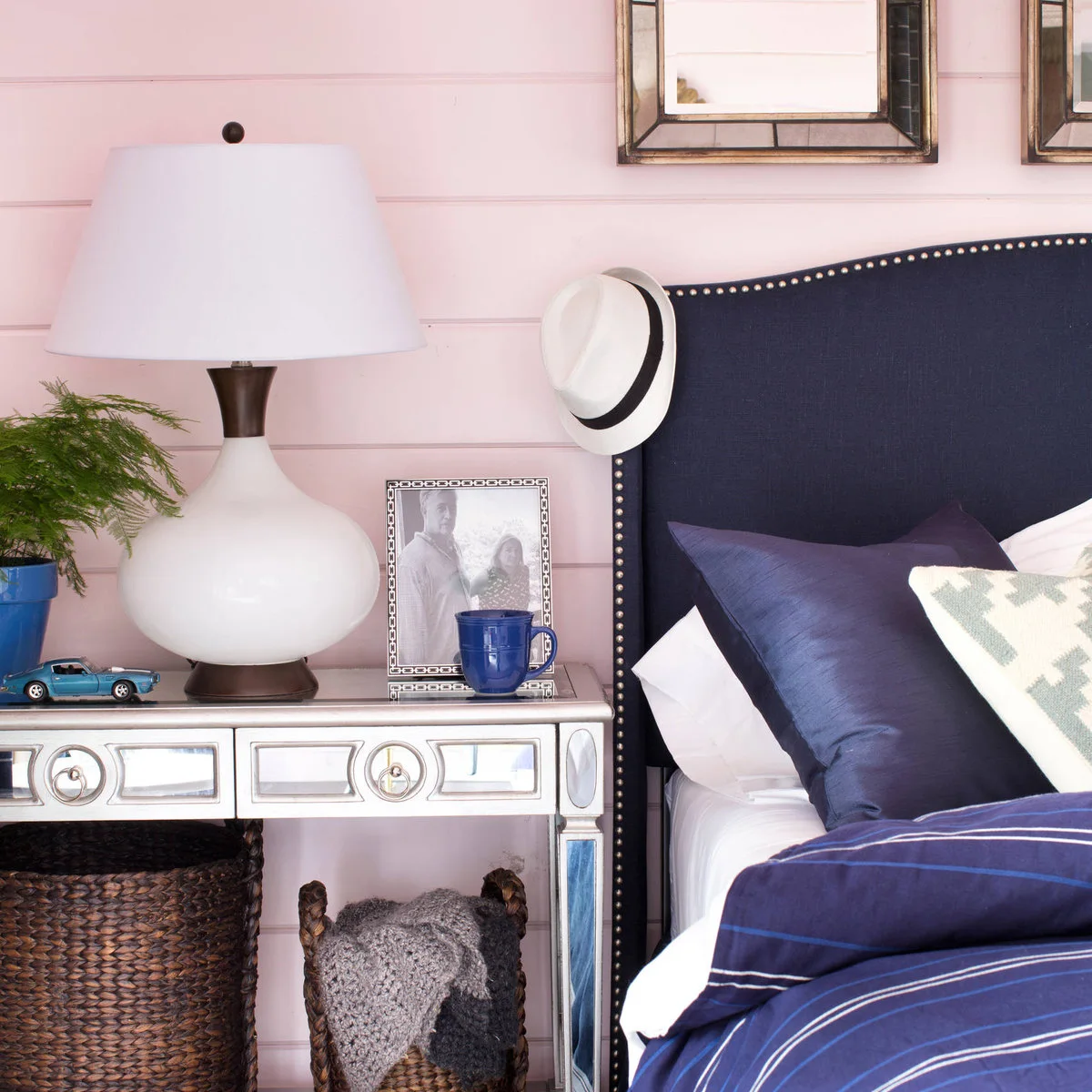 Bedside view of a pale pink paneled wall, navy studded headboard, white lamp and mirrored bedside table