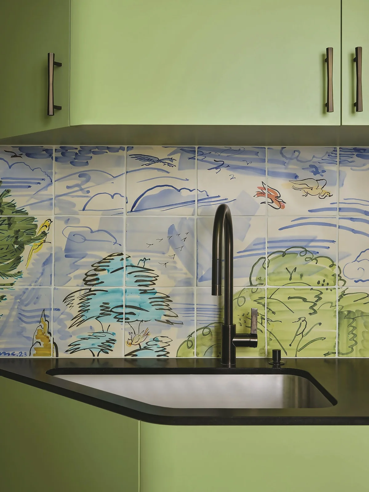 Blue-and-green painted backsplash tiles sit above a dark sink and counter beneath pale green cabinet doors