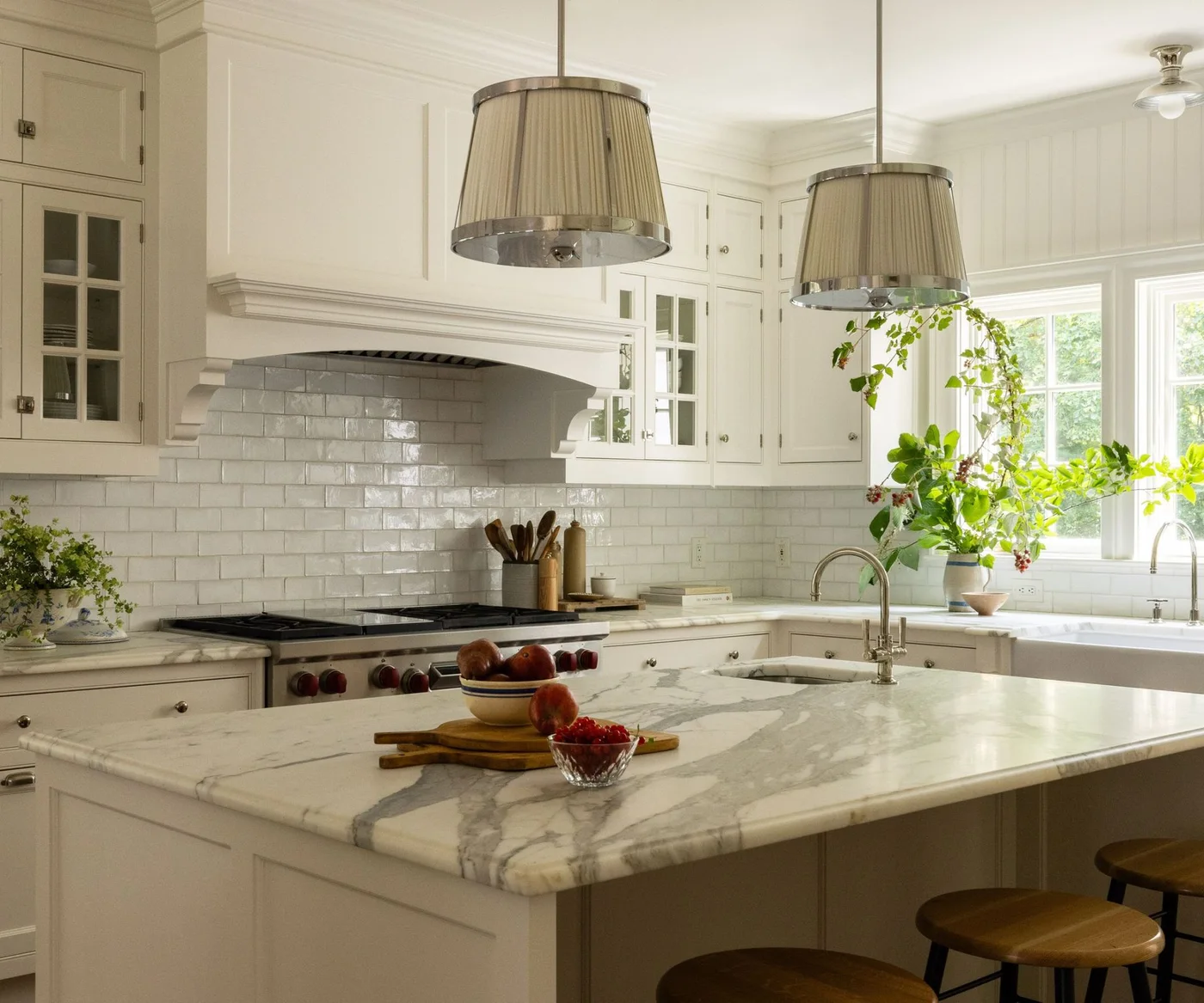 A white kitchen has a detailed cooker hood, glazed upper doors, glossy rectangular tiles, two pleated pendants and a veined island with a small sink.