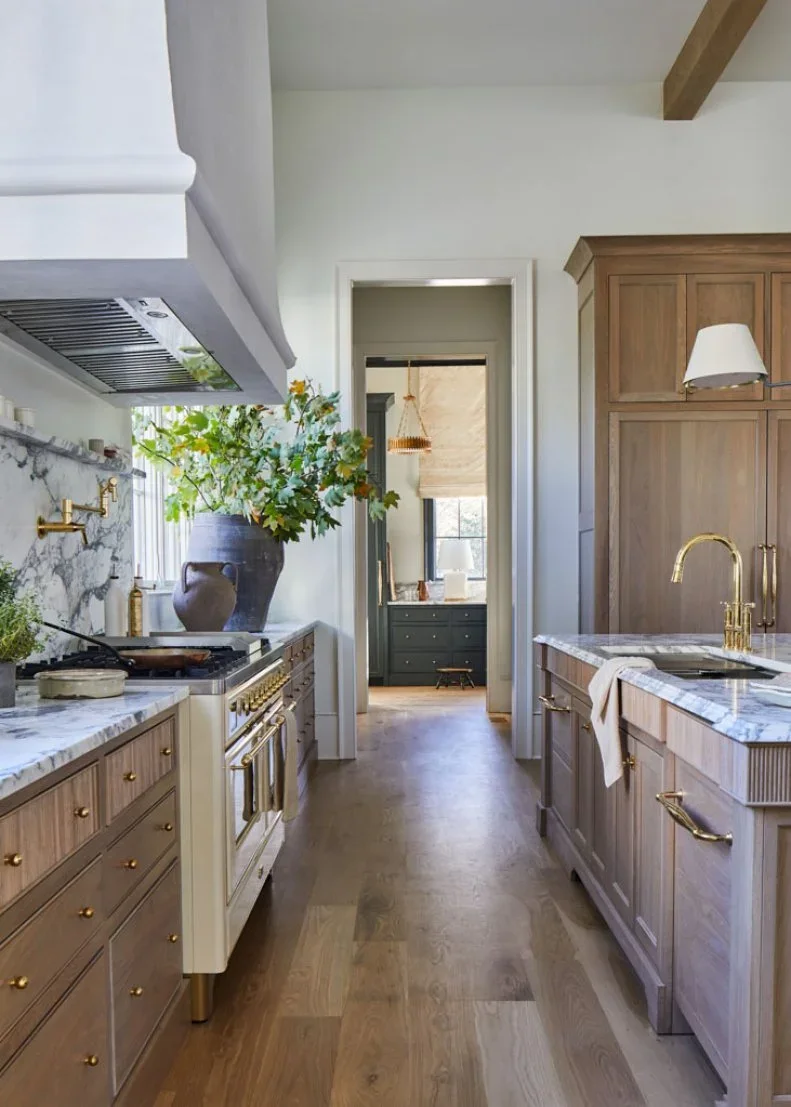 White oak drawers face an island sink across a clear aisle, with a range, pale hood and an open doorway to a darker pantry.