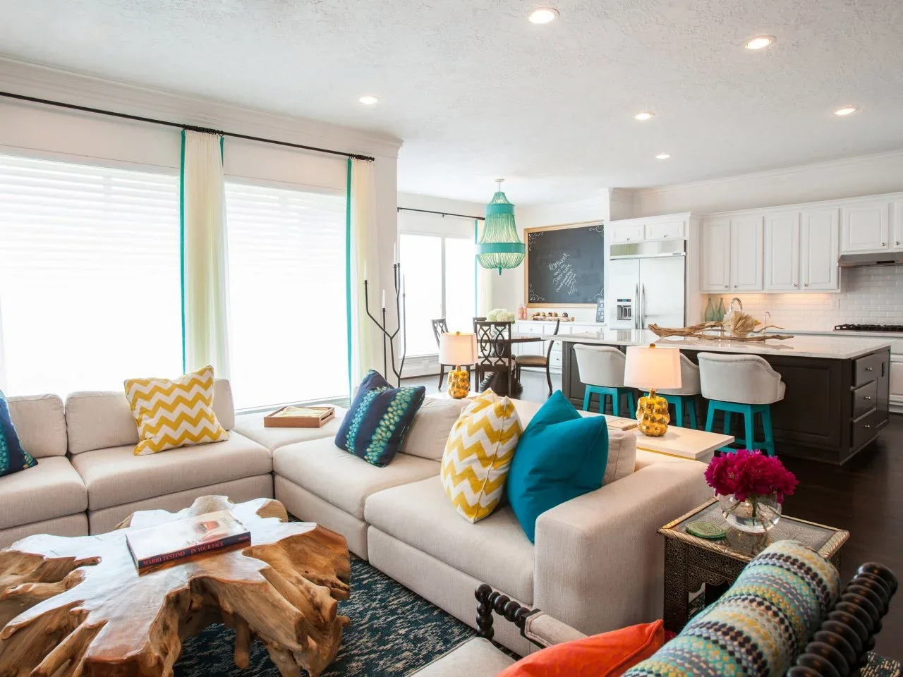 A pale sectional with teal and yellow cushions sits beside an open white kitchen with teal-legged stools and a teal dining pendant.