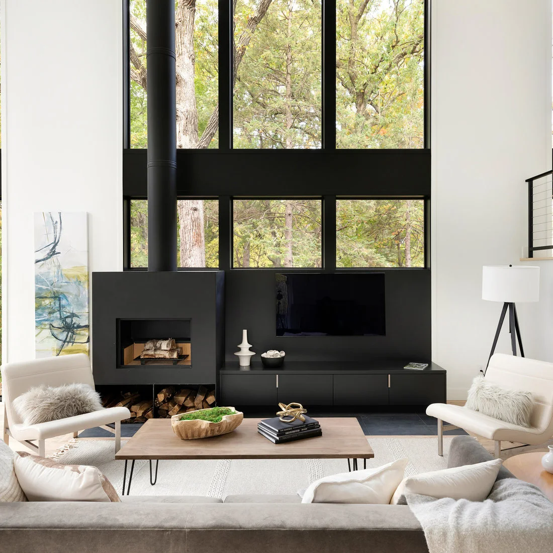 A black fireplace and flue stand beside matching low media cabinets beneath tall black-framed windows, opposite pale chairs and a simple wood coffee table.