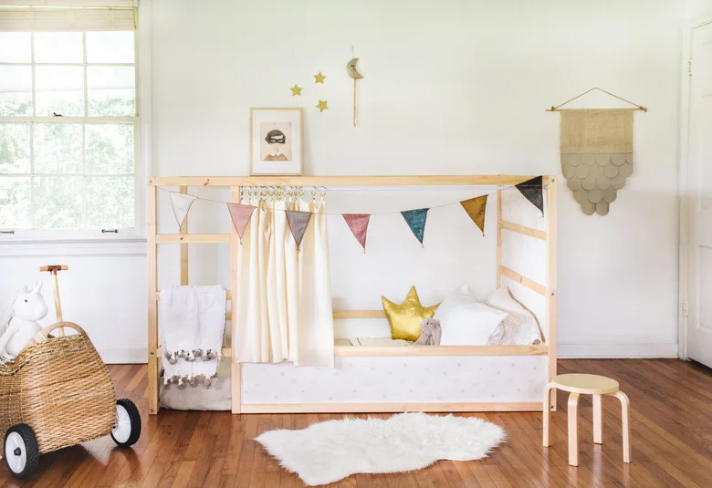 A pale wood bed with an open upper frame and small bunting stands against a white wall beside a woven toy pram and a small stool