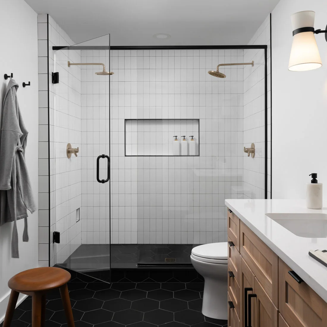 A clear black-framed shower has white vertical tiles, two warm-metal shower heads, a central niche and a dark hexagonal floor beside a wood vanity.