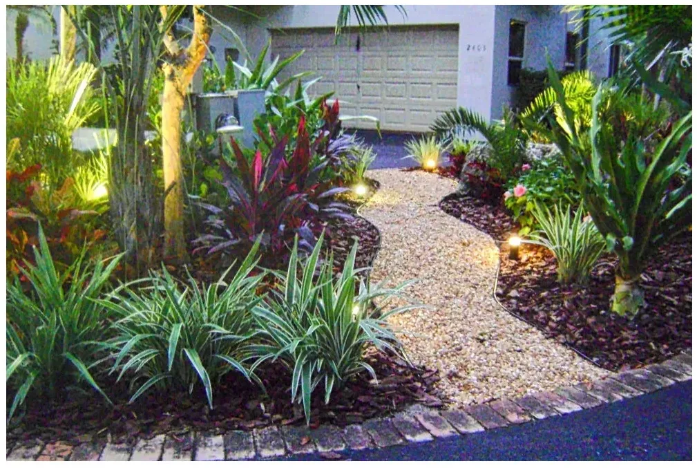 A curved gravel walk between mulched tropical beds, low striped leaves, taller palms and small path lights near a garage.