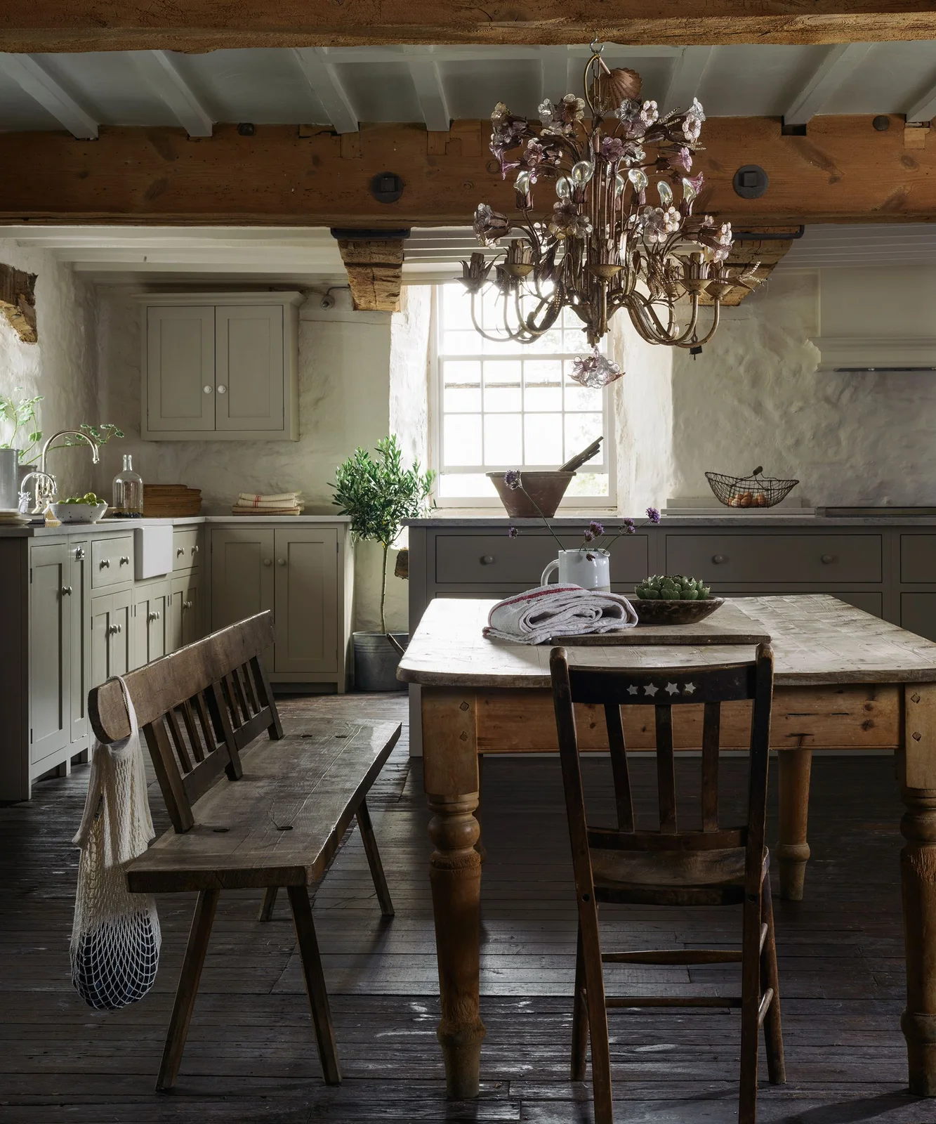 Gray-green kitchen cabinets surround a wooden table, bench and chairs below exposed beams and a decorative chandelier beside a white apron-front sink
