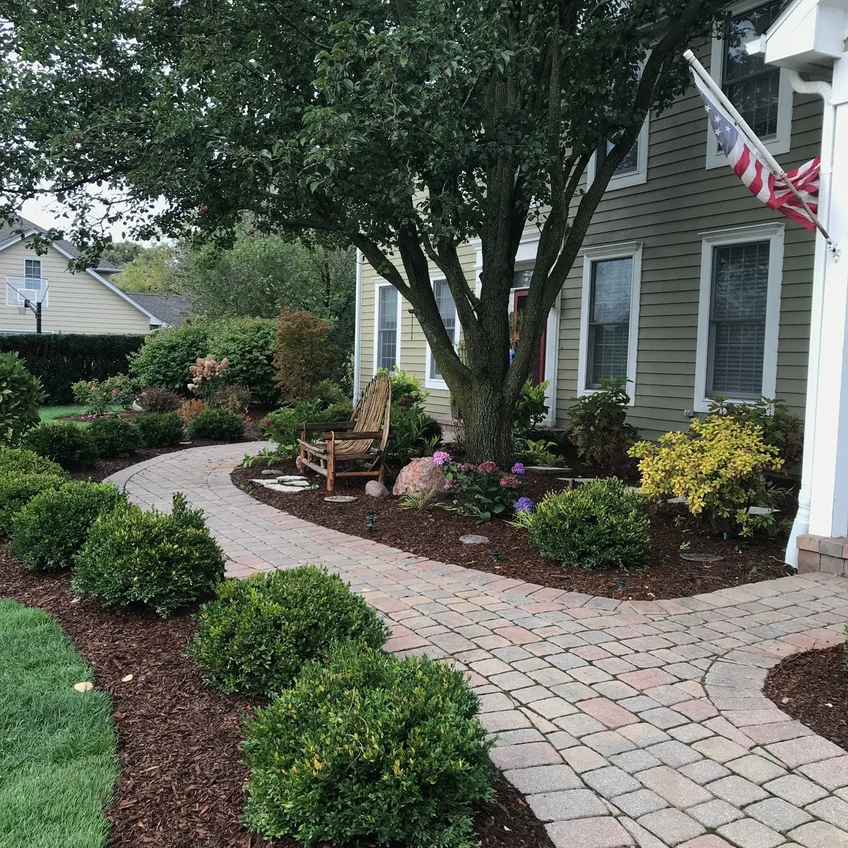 A curved paved front walk beside a mulched bed, with a wood chair under a mature tree and grouped shrubs near the house.