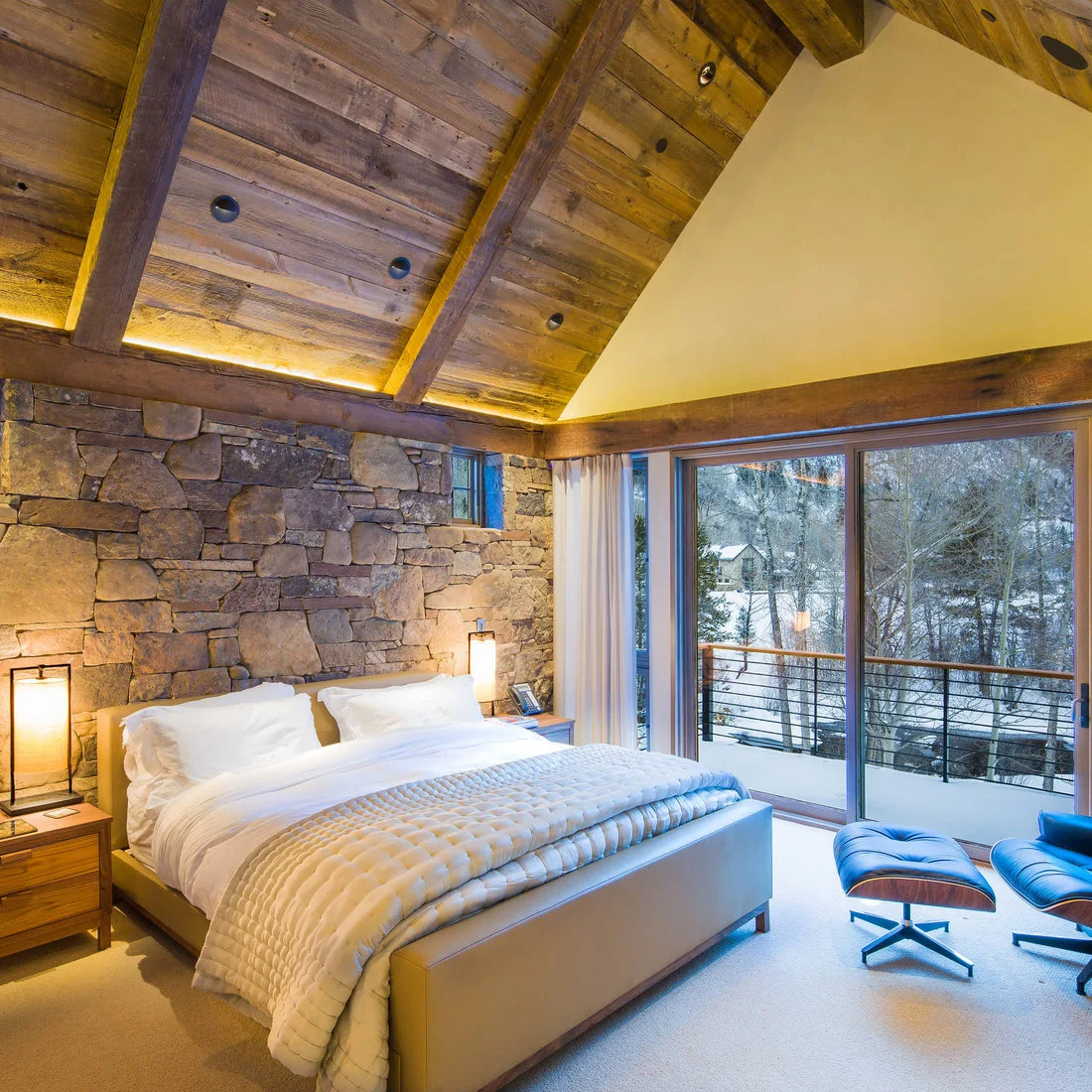 A pale bed and timber bedside drawers against a rough stone wall below a timber ceiling