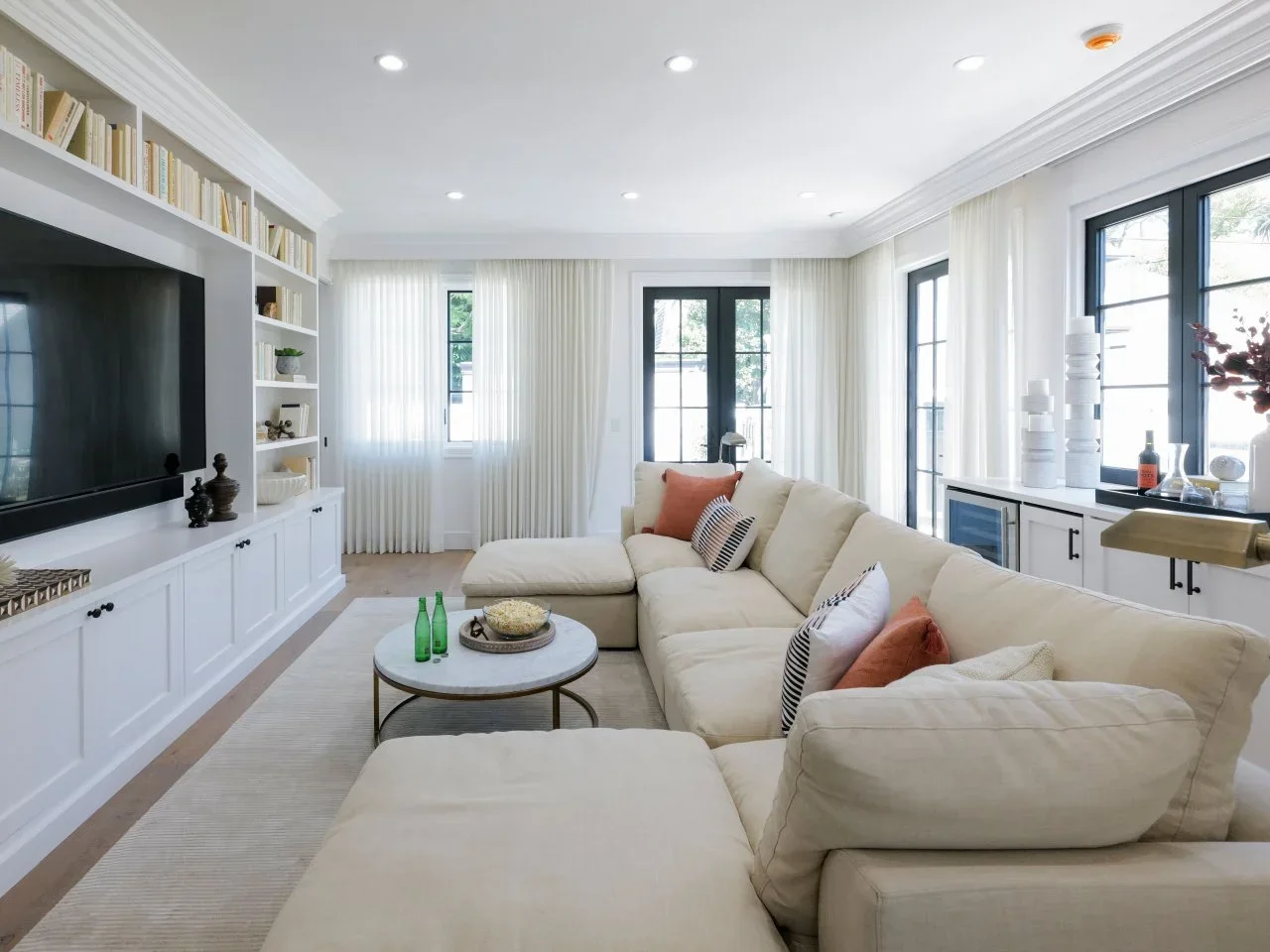 A cream sectional faces white built-in television storage beside black-framed windows and light curtains.