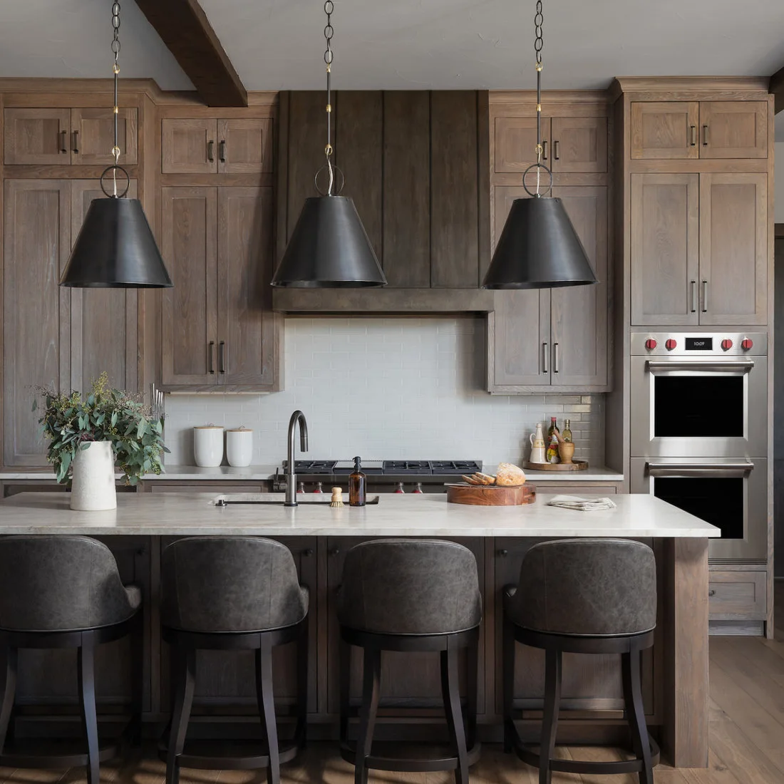 Wood cabinets and a dark hood behind a pale island with black pendants and upholstered stools