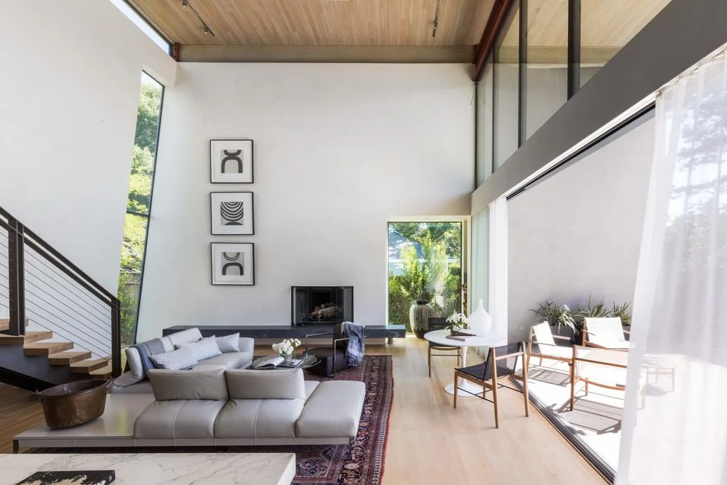 Pale sectional seating and light-framed chairs sit beneath a tall white wall with three small artworks, broad glazing, a wood ceiling and a nearby staircase