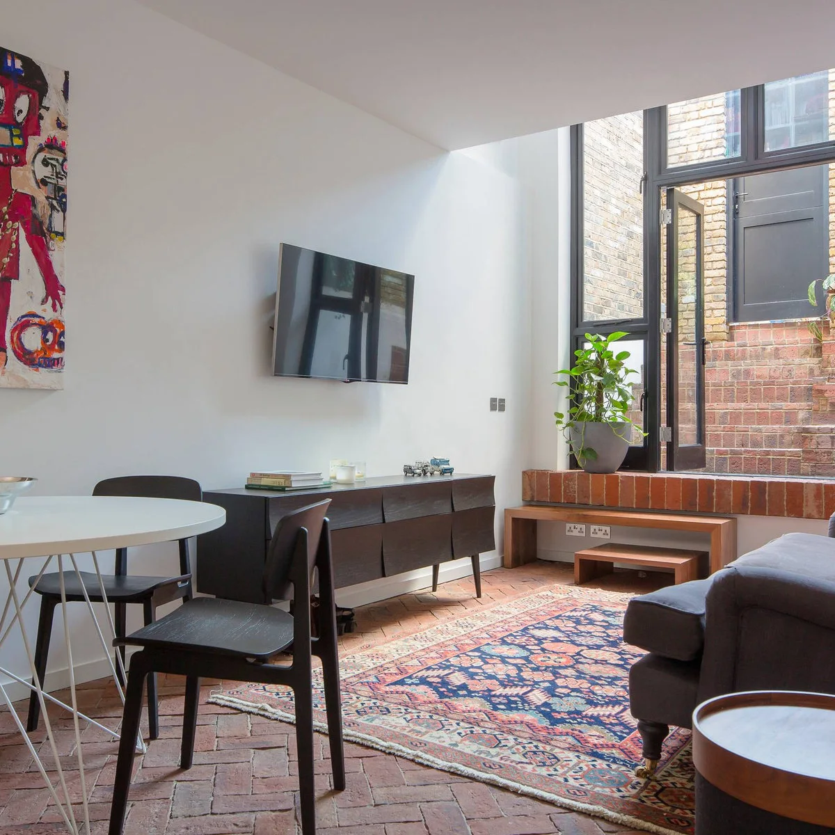 A compact brick-floored living room has a round white dining table, black chairs, a gray sofa and a wall television above a dark low cabinet.