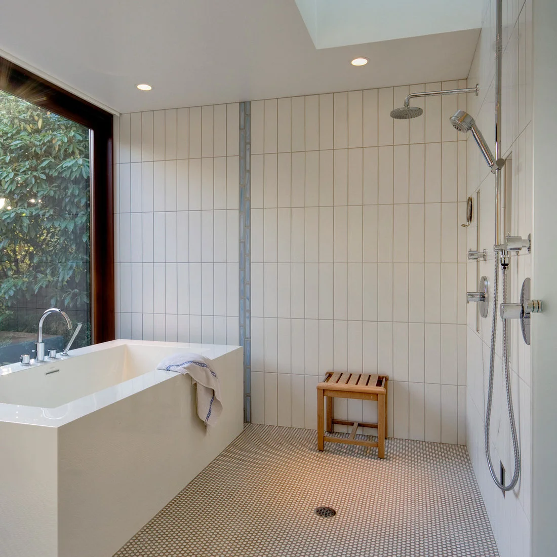 Small pale round floor tiles with contrasting grout run below a white tub and an open shower with a timber stool.