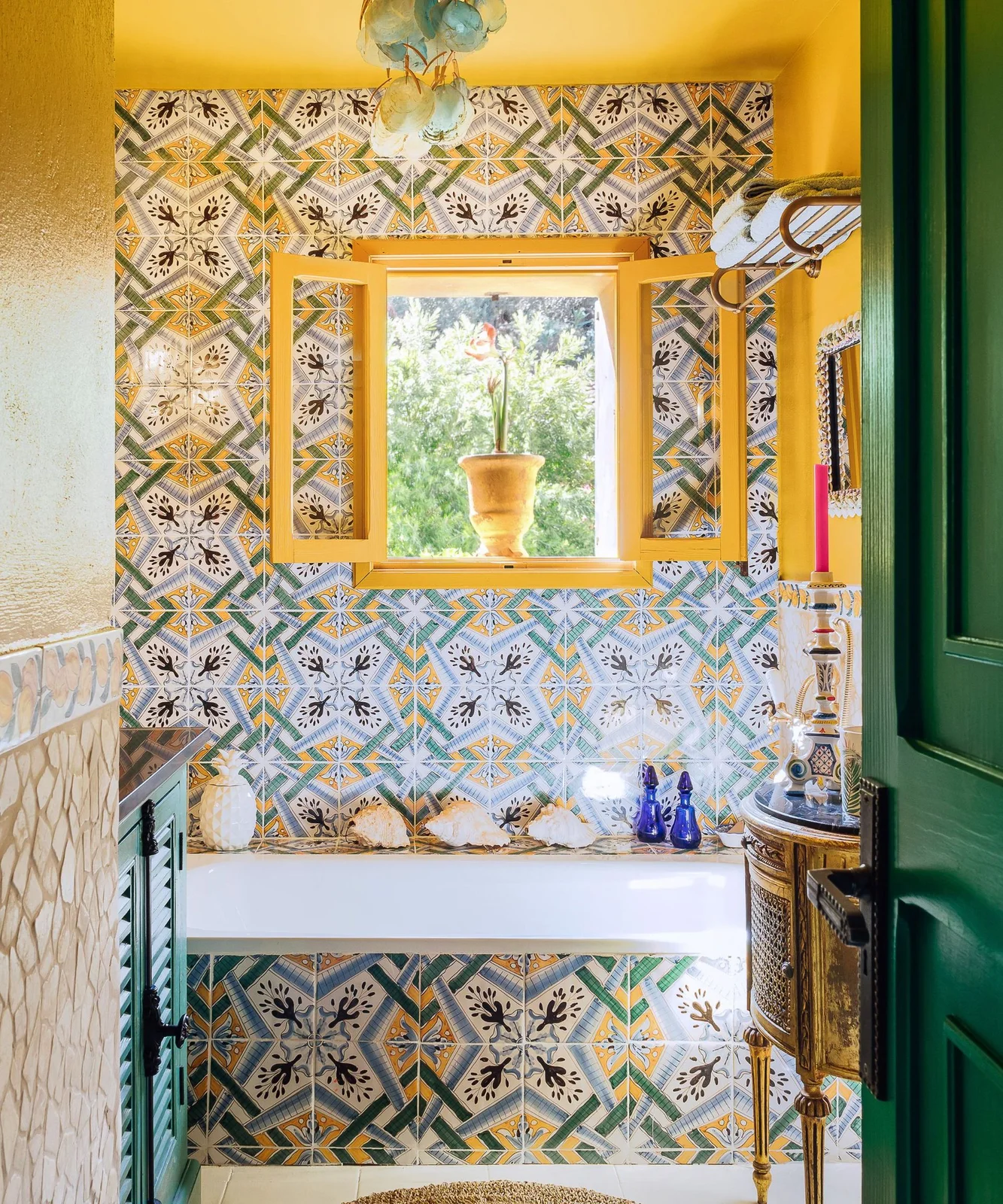 A small bathroom has yellow walls and window trim, green doors and cabinetry, multicolored patterned tiles around the bath and decorative objects on the ledge.