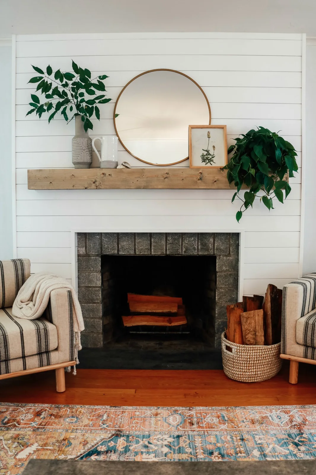 A white shiplap fireplace wall has a simple thick wood mantel, dark brick opening, round mirror, leafy arrangements and striped chairs at either side.