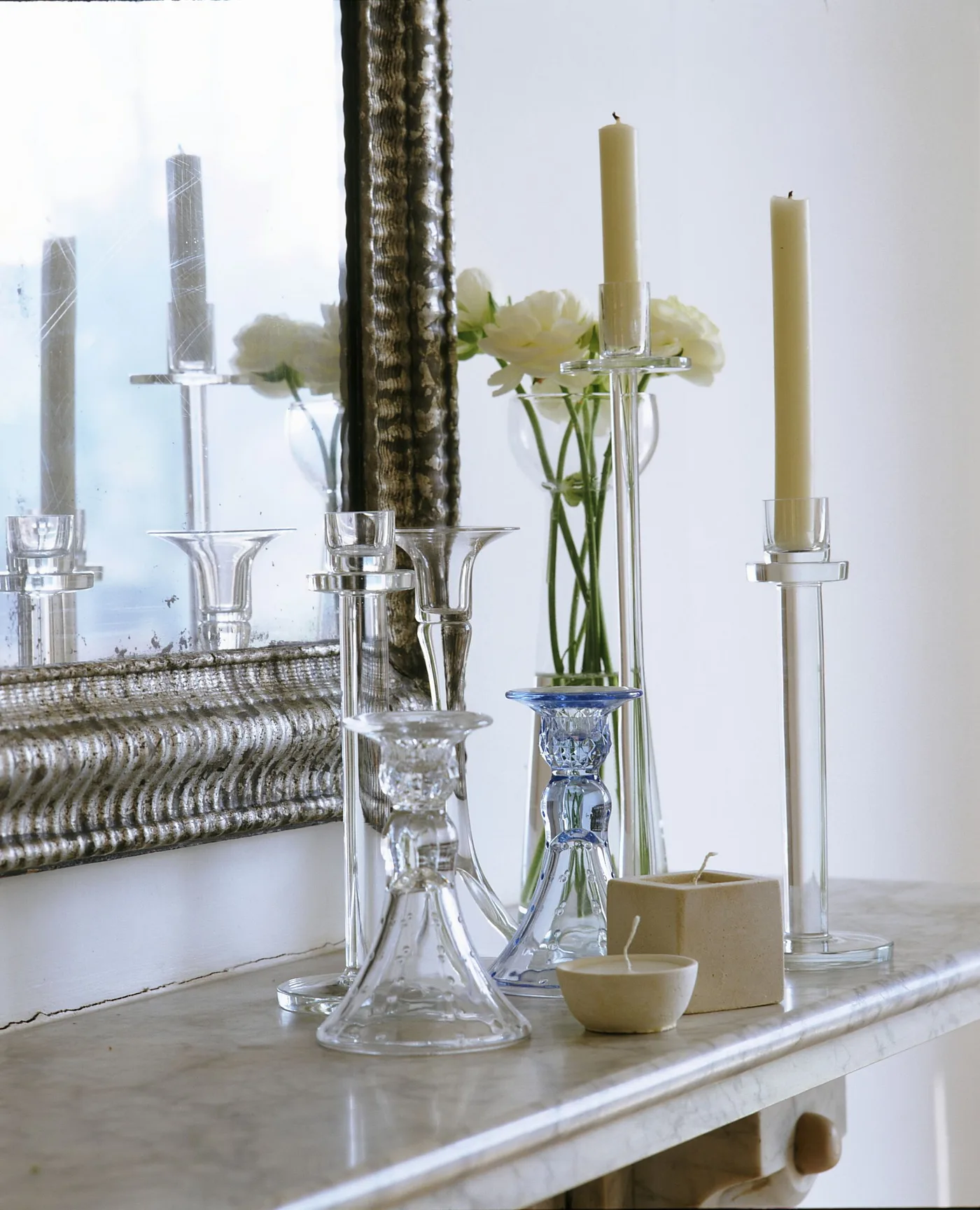 Pale mantel with reflective mirror, clear and blue-toned glass candle holders, pale candles and flower vase