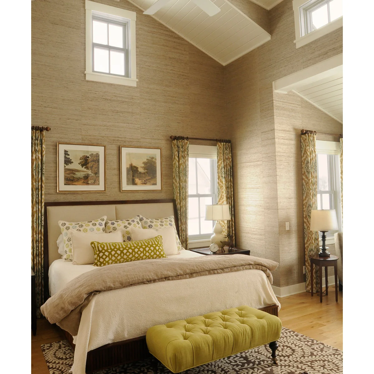 A brown and cream floral rug sits beneath a pale bed and a green tufted bench in a tall bedroom.