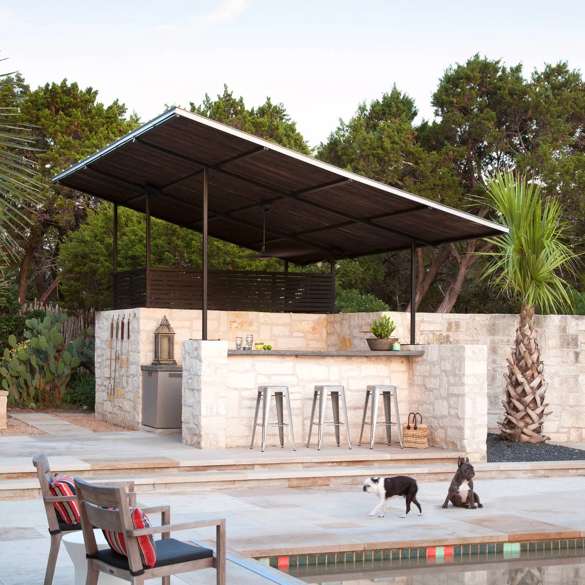 Small stone-faced outdoor bar with three metal stools beneath a single-slope roof and timber screening