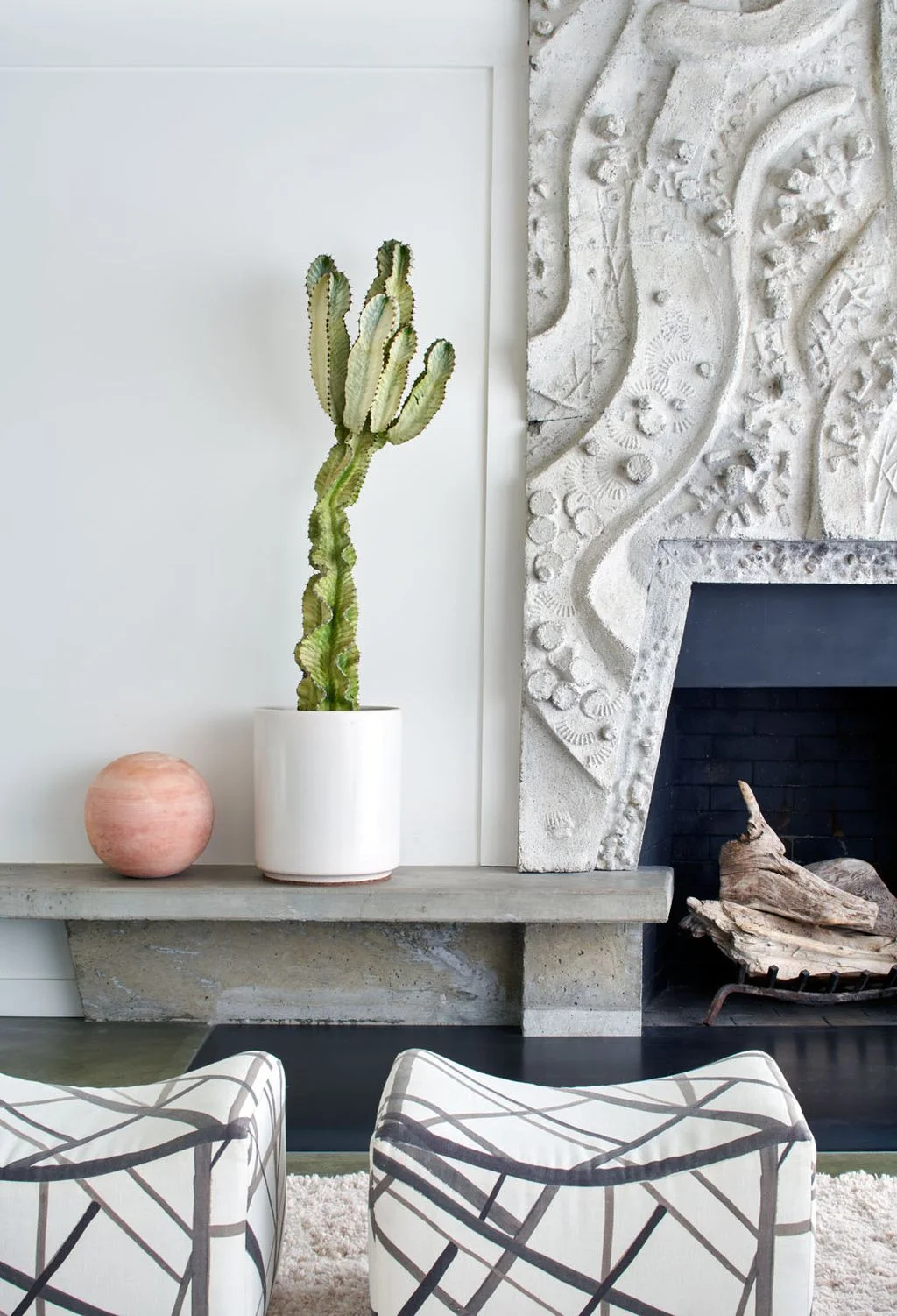 A pale sculptural relief surrounds a dark angled fireplace opening beside a gray low shelf, a tall cactus and two patterned upholstered stools