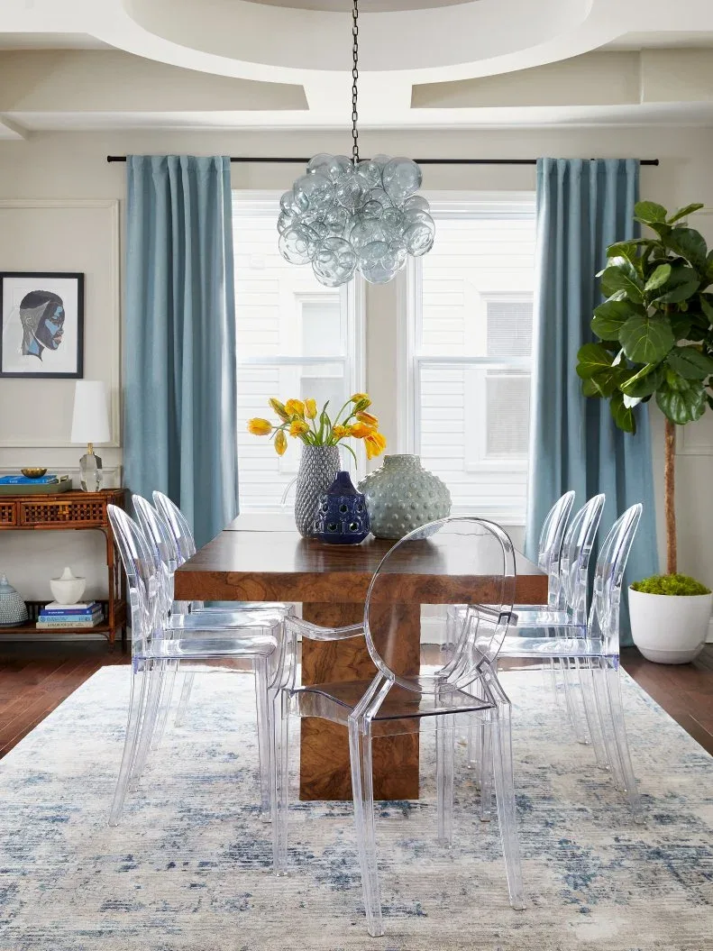 Plain blue curtains around a bright dining-room window, with a pale blue globe-cluster light, wooden table, transparent chairs, and large leafy plant.