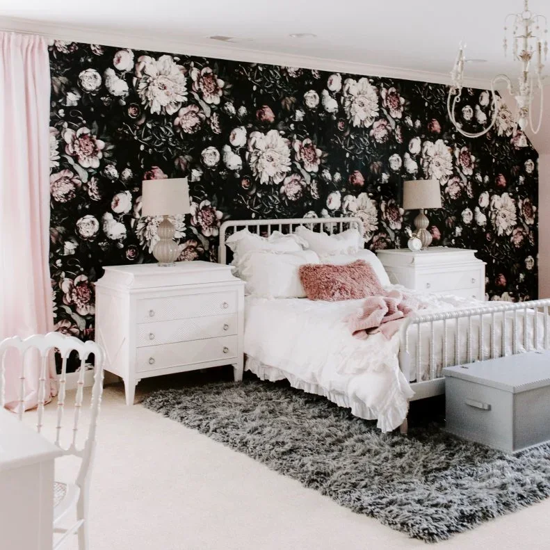 Pink and white flowers cover black bedroom wallpaper behind a white metal bed and matching drawer chests.
