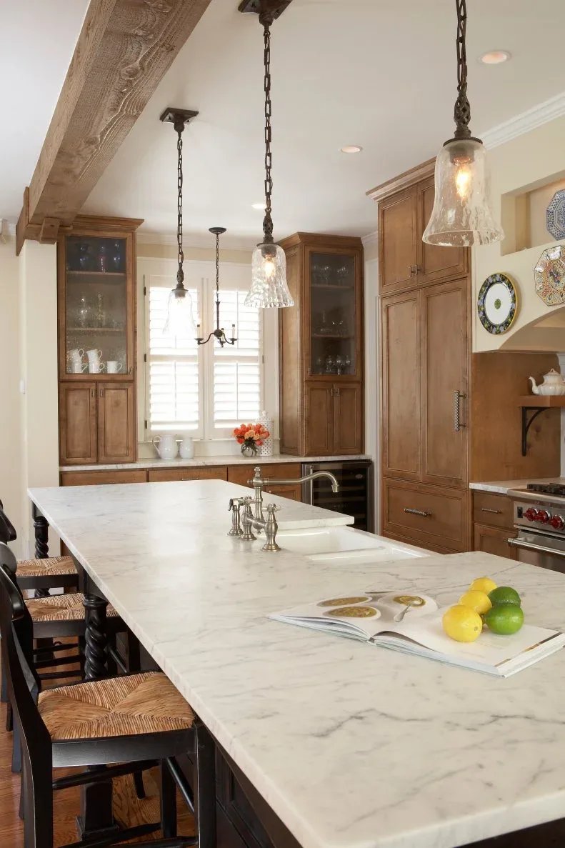Warm timber kitchen cabinets and textured glass doors sit behind a pale island beneath hanging glass lights.