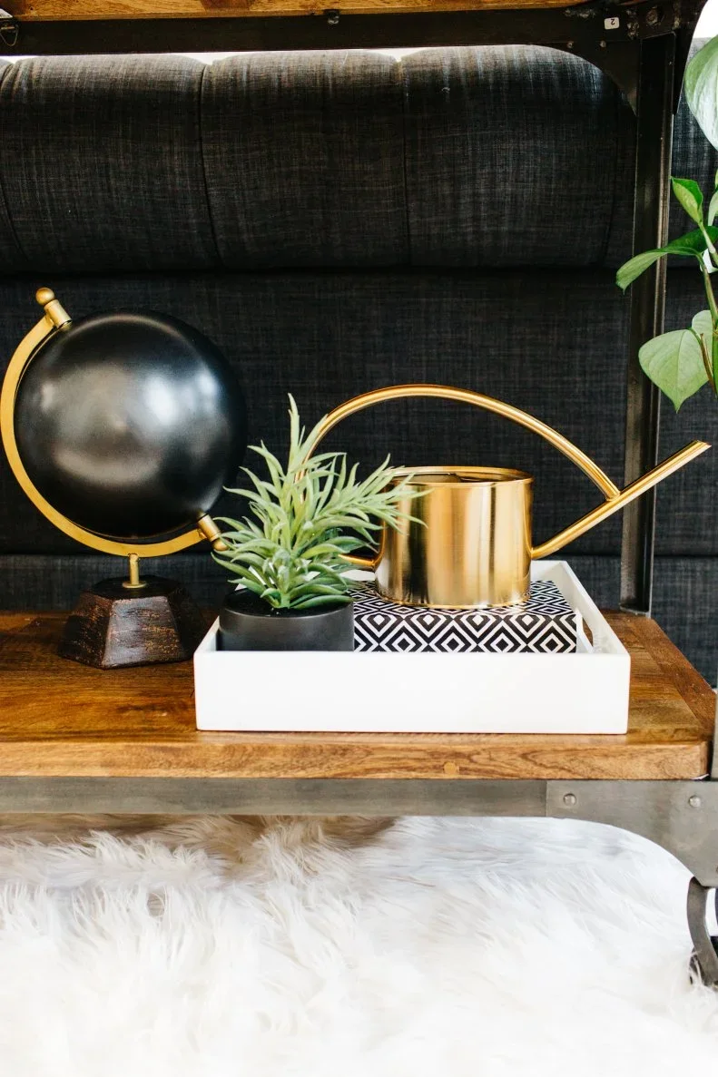 A white tray holds a small plant and gold-toned watering can beside a black globe on a sofa-console shelf.