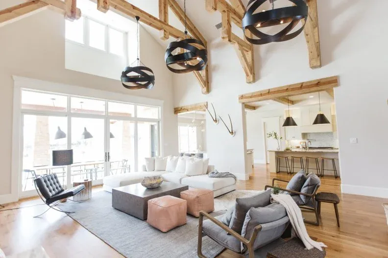 High country room with exposed timber trusses, black ring pendants, white sofa, gray wood-framed chairs and two pale square stools
