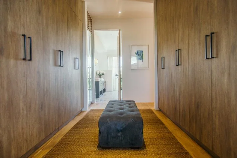 Timber closet doors face each other across a narrow dressing room with a dark tufted bench and yellow-toned rug.