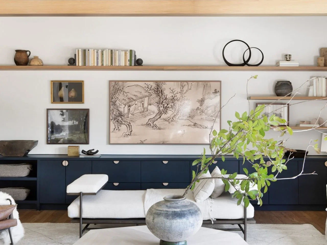 A long timber shelf and shorter shelves sit above navy fitted cupboards, with framed art between them and pale seating in front.