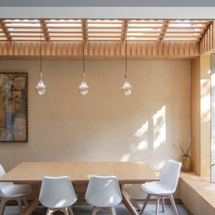 A pale wood table and white chairs sit below a slatted rooflight and three clear pendant bulbs beside a low wood window seat.