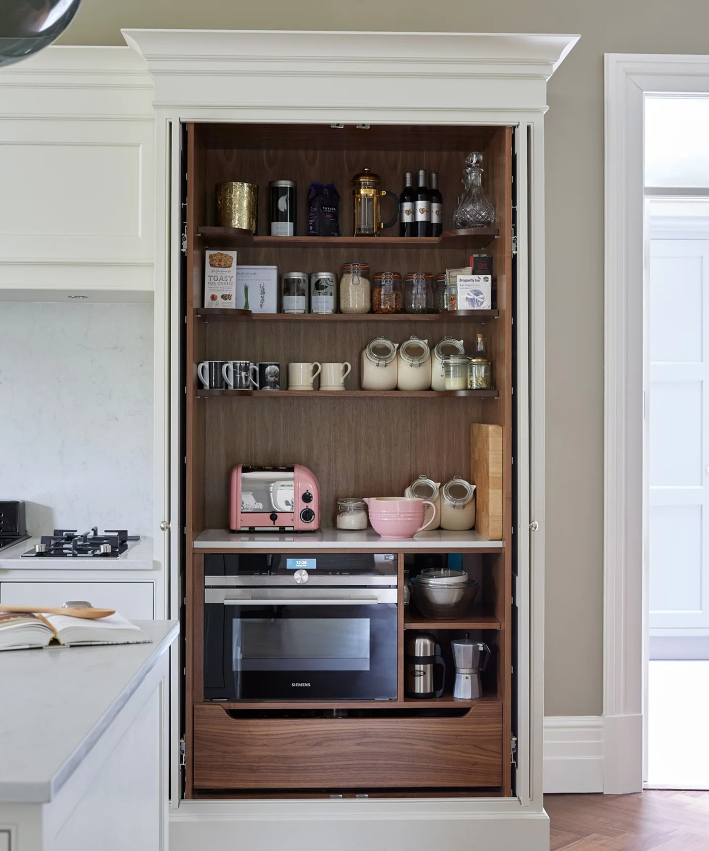 Tall pale paneled kitchen cupboard opened to reveal wooden shelves, food jars, cups and built-in appliance storage.