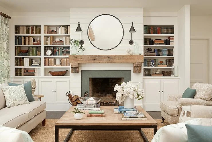 A white shiplap fireplace wall has a wood mantel, round black-framed mirror, paired wall lights and fitted bookcases above lower cupboards.
