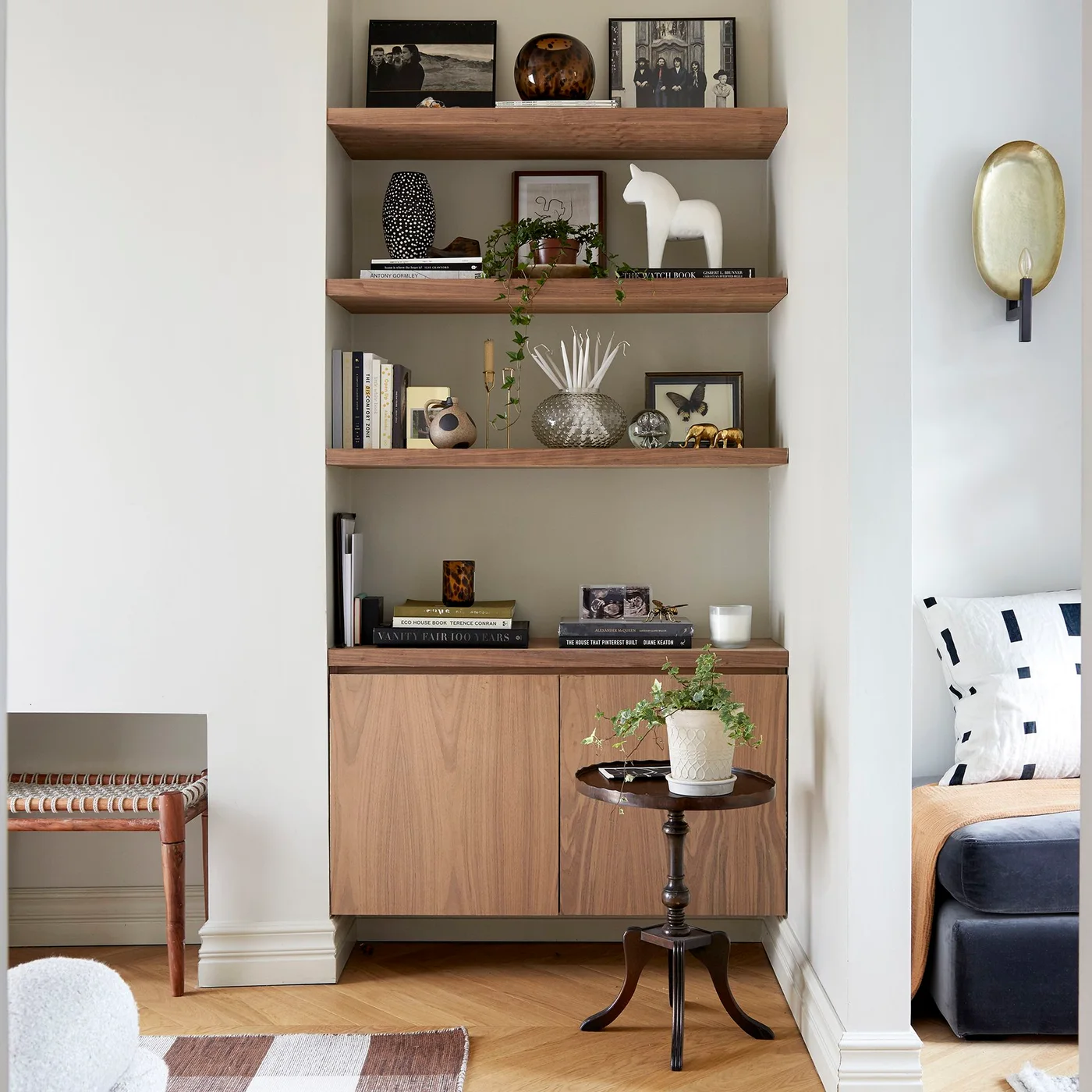 Timber shelves and a closed cupboard fill a pale sitting-room alcove beside a dark seat and a small round table.