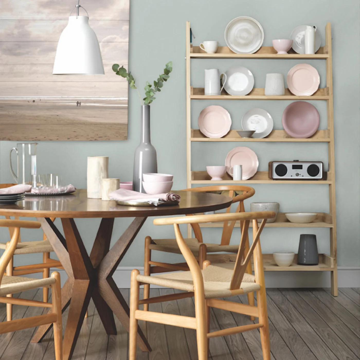 Pale green walls sit behind open wooden plate shelves beside a round dark table, wooden chairs and a pale pendant