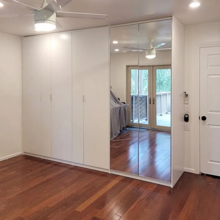 Wall of white PAX doors with two mirrored panels reflecting a bright bedroom and terrace doors