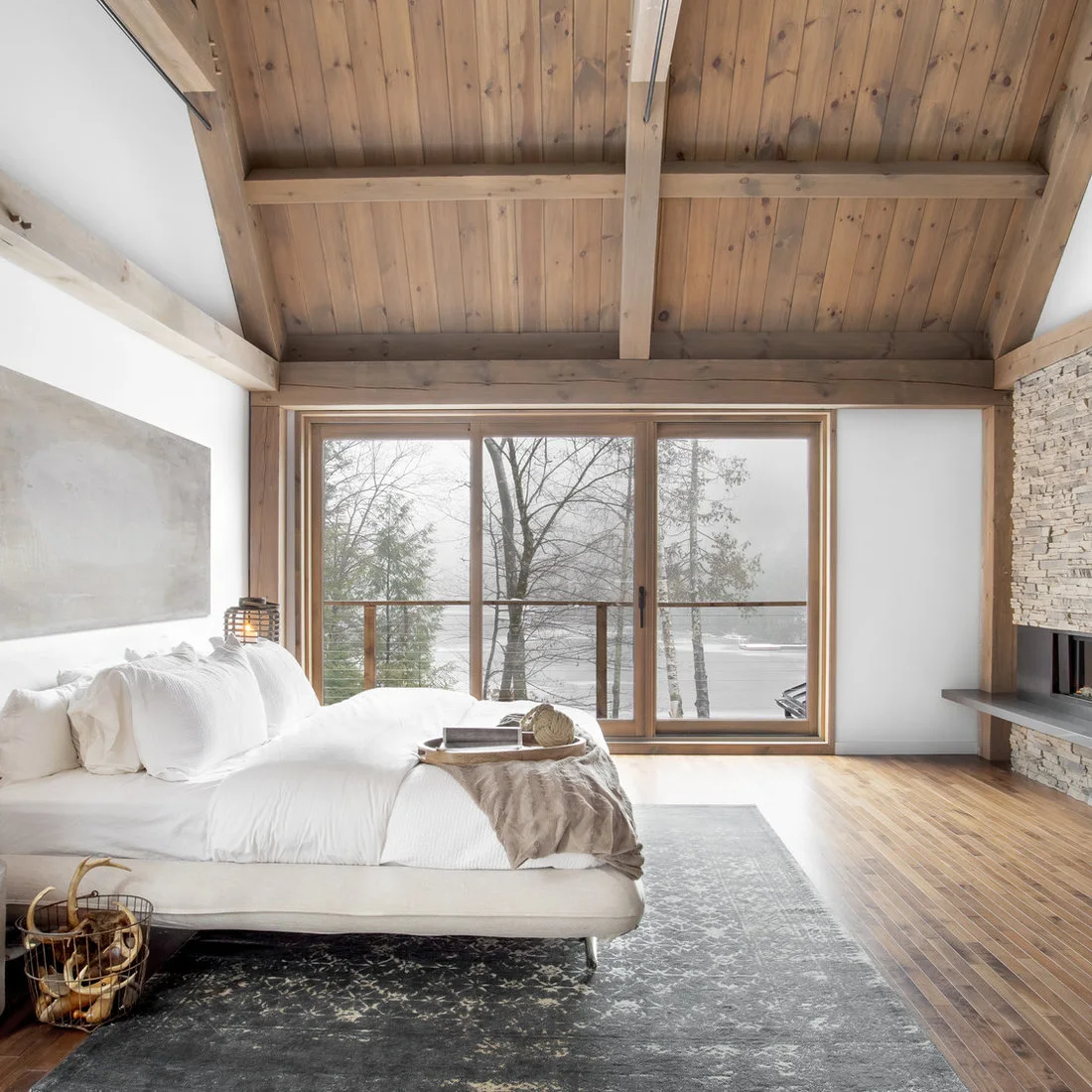 A low pale bed on a dark rug beneath exposed timber beams beside a stone fireplace
