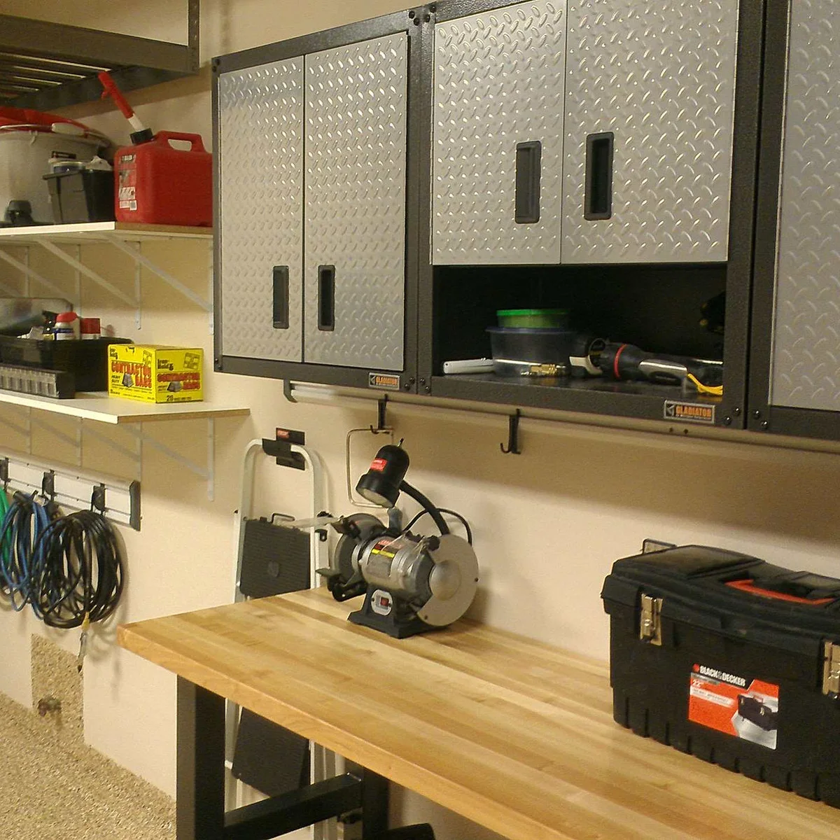 Wood-topped garage bench with a small bench grinder, metal wall cabinets, a toolbox and coiled cables nearby.
