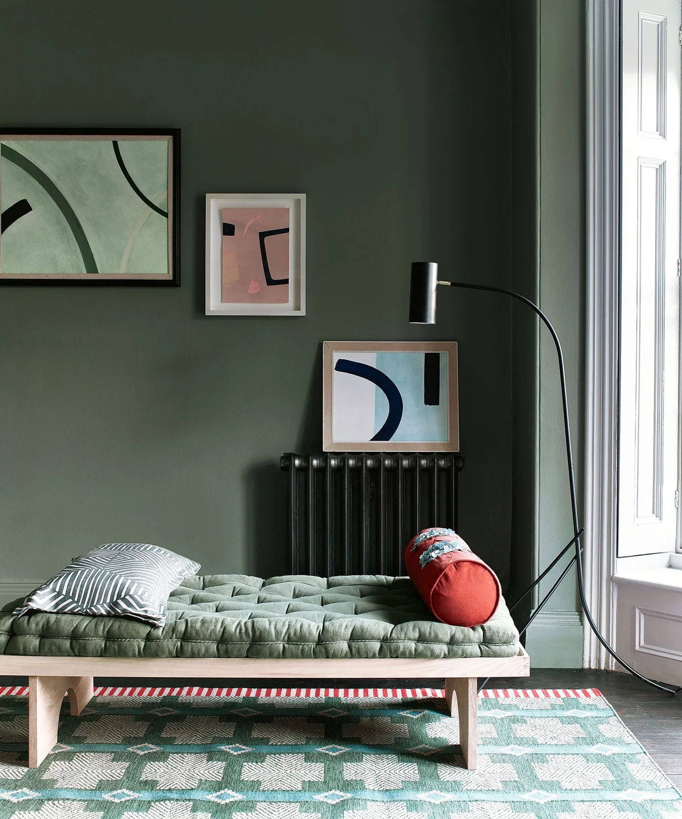 A tufted green daybed with a red bolster sits against muted green walls beside a black floor lamp and framed abstract pictures