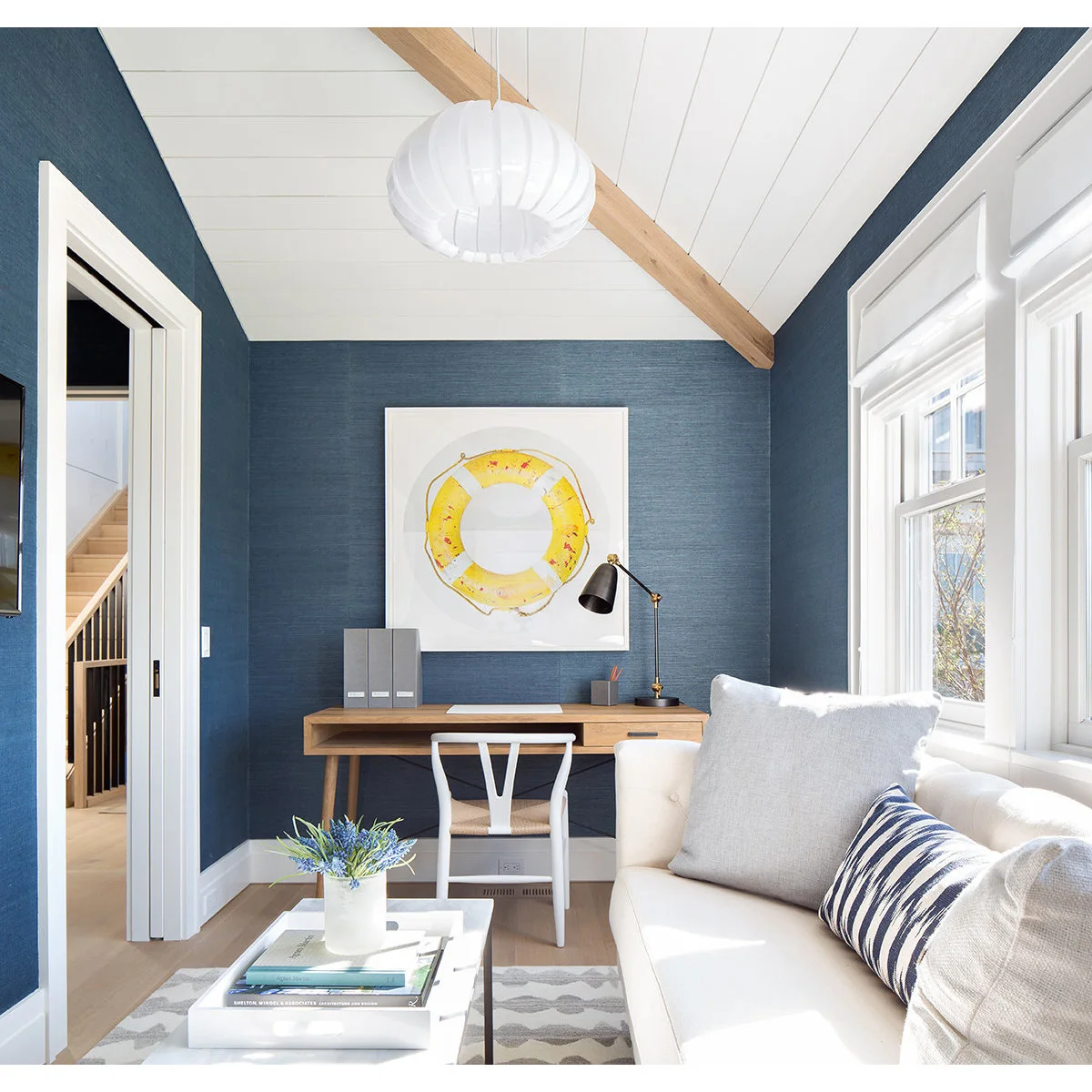 A slim wood desk against a blue end wall beside a pale sofa and a large window.